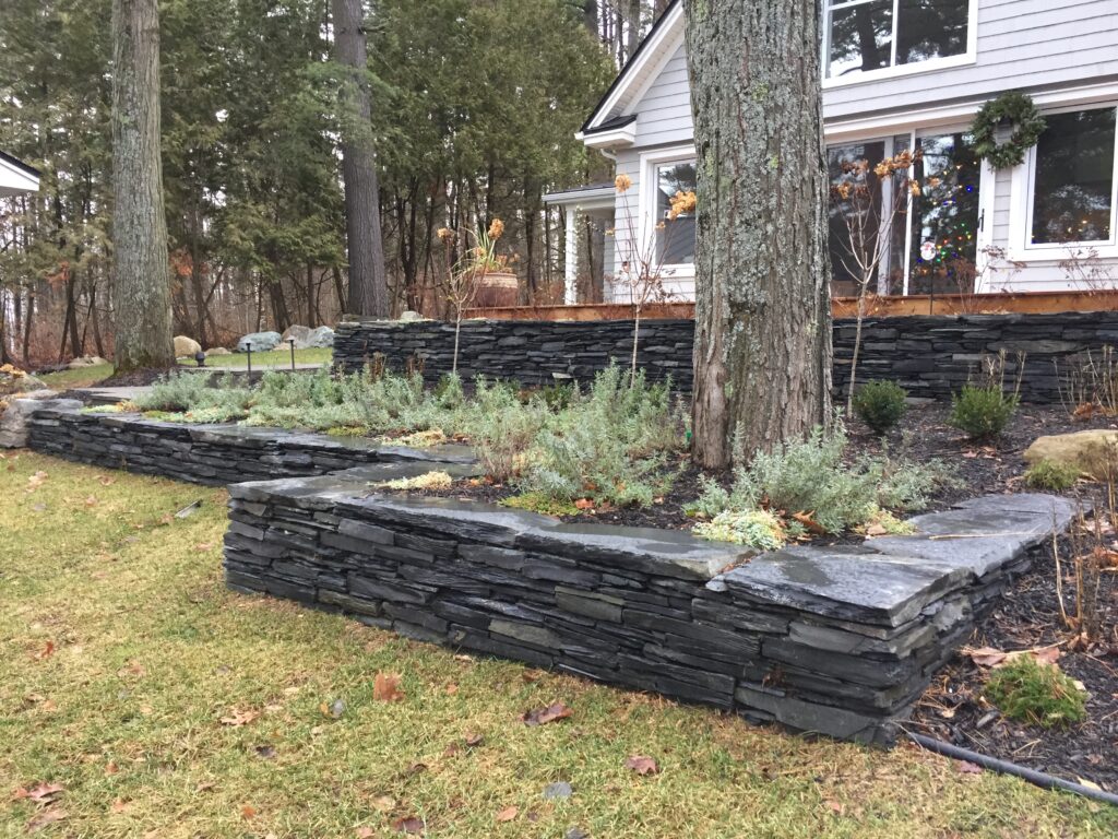 Muret en pierre entourant et surélevant des arbres et plantes en bordure d'une maison
